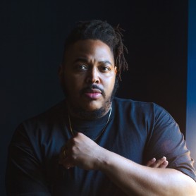 A picture of a man named Darius Clark Monroe. He is wearing a black quarter sleeve t-shirt with his neck adorned with a gold chain. He has one of his hands across his body touching the chain, as he stares with a serious expression at the camera ahead. He has black locs a mustache and sparse beard.