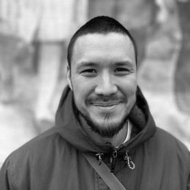 A black-and-white picture of a man named Aidan Un. He is standing in front of a blurred, textured background with light and dark areas. He has a short haircut and a neatly trimmed beard. He is wearing a dark hooded jacket with a strap across the chest. He is smiling towards the camera, with his eyes slightly squinted.