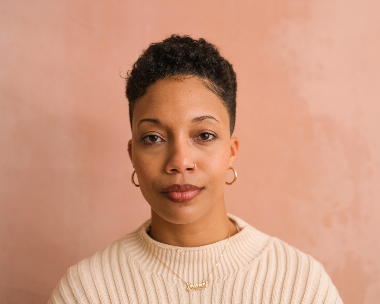 A headshot of Dessane Lopez Cassell. She is wearing a white shirt and in front of a salmon colored backdrop, looking directly at the camera.