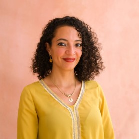 A headshot of Nehad Khader, she is a Palestinian woman. She is wearing a flowy yellow top. She has curly hair. She is smiling softly at the camera.