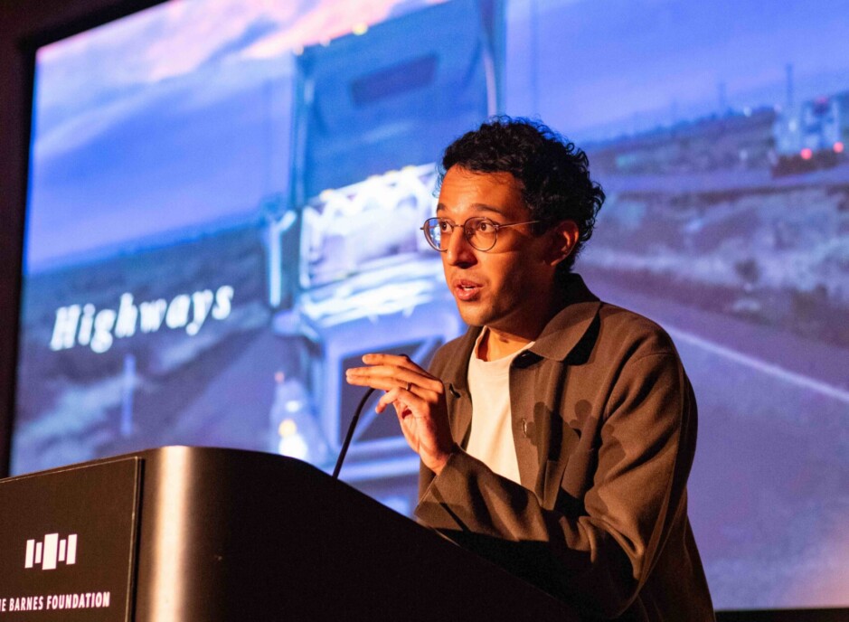 A photo of a person speaking into a microphone in front of a screen.