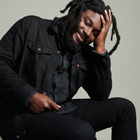 A photo of Jason Reynolds shows him wearing all black and smiling brightly towards the camera while resting his head in his left hand.