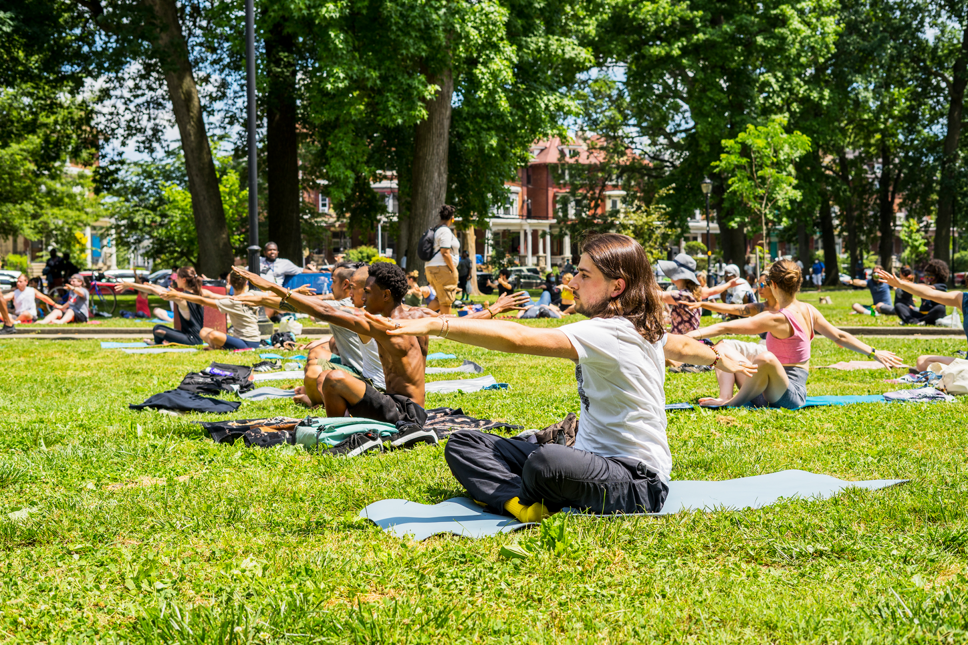 Outdoor photo of a group of people sitting on a large grassy area. They sit on yoga mats and stretch their hands out in front of them.