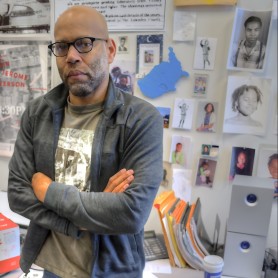 A photo shows a portrait of Kevin Jerome Everson standing inside his workspace with his arms folded and looking directly at the camera.