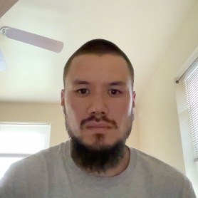 Photo of photograpoher Aidan Un shows him sitting in a room with windows behind him. He wears a grey shirt and has a long black beard.