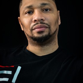 Photo of photographer Daniel Jackson shows him standing against a black. background and wearing a black t-shirt that blends into the background. His facial hair is cropped to his face and he looks into the camera.