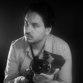 A hazy black and white picture of a person named Suneil Sanzgiri. He is sitting on something and against a black background and side lit. His face is turned towards the left and he is holding a camera looking off to the side in thought. He has a light mustache and sparse beard and is wearing an open button down top.