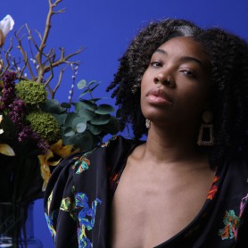 Image of Kerrin Lyons shos her posing with a large floral bouquet filled with various plants and flowers. She wears a black embroidered top with a deep v-neck and tilts her head slightly up as she looks into the camera.