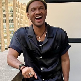 Photo of Michael Moody shows him standing outside on a balcony. He wears a green beanie on his head and a black button-down shirt. He leans forward on his forearm and laughs.