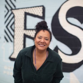 A picture of a woman named dream hampton. She is outside in front of a blue painted wall with two letters, slightly leaned forward and showcasing a radiant grin. She is wearing a dark button down top with a v-neck line, and her neck is adorned with a blue beaded necklace. She has her curly hair styled atop her head in a bun.