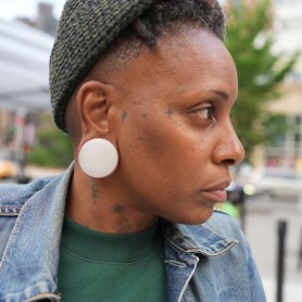 Headshot of the artist Lynneé Denise shows her wearing a jean jacket and green knitted hat. She wears large white ear pluggs in her stretches ears and has several small tattoos of stars and dots scattered on her neck and face.