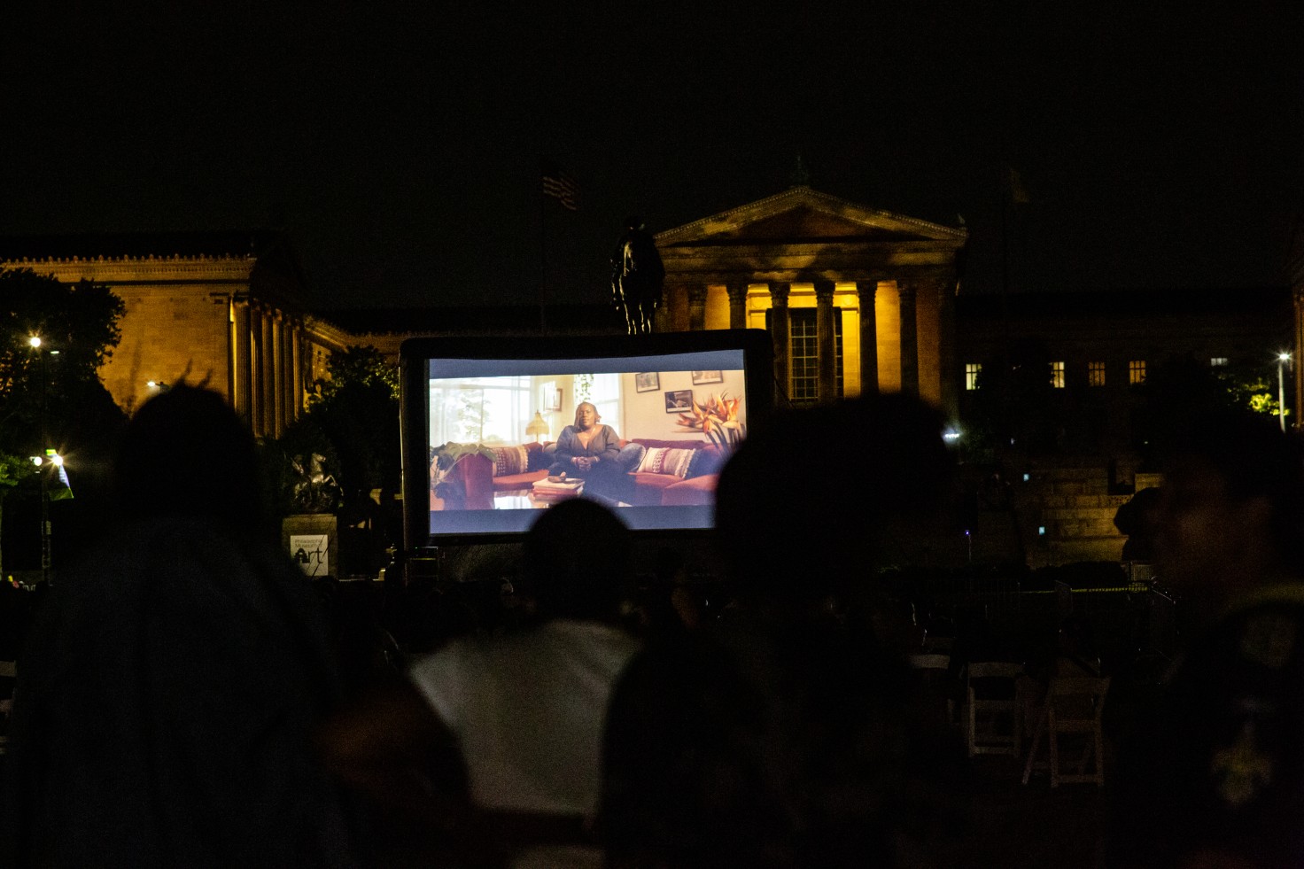 A photo a film screening taking place outside of the Philadelphia Museum of Art. It is dark outside. The large blow up screen has an image of a Black woman on it. The art museum is illuminated in the background.