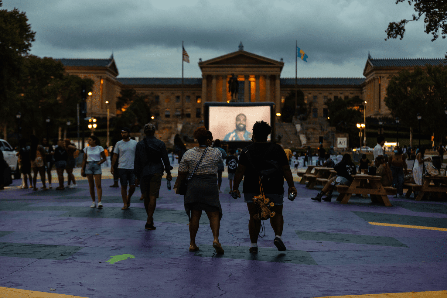 A photo of a film screening taking place outside of the Philadelphia Museum of Art. Two people are in the foreground walking towards the screen.