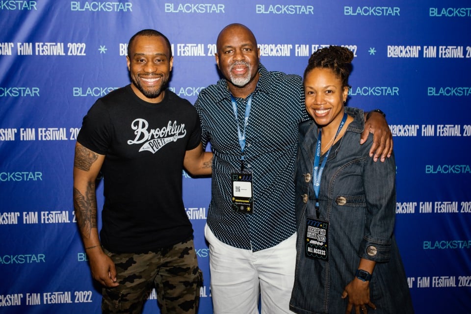 A photo of Byron Hurt, Marc Lamont Hill, one other person posing in front of the step-and-repeat. They are all smiling.