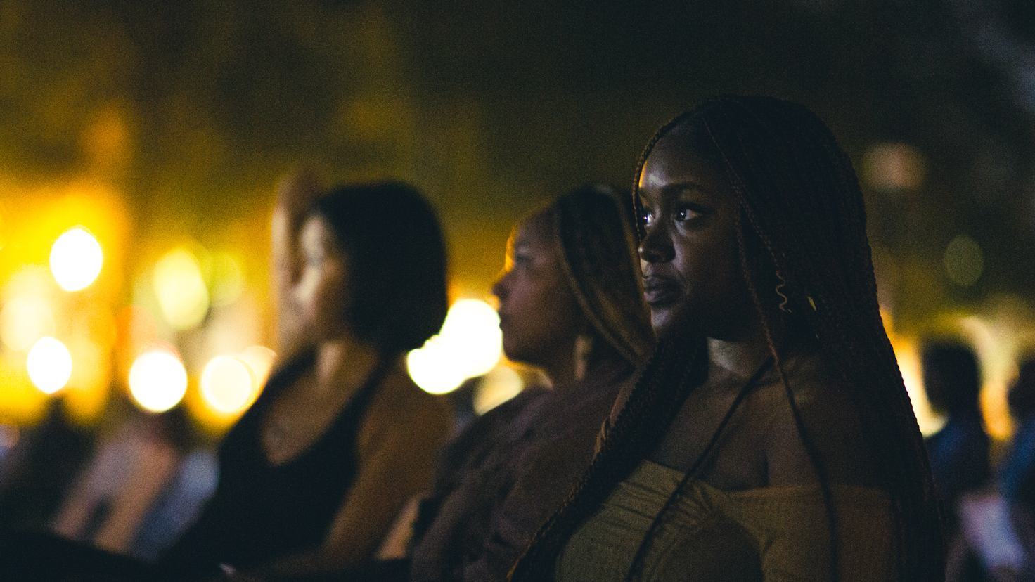 A photo of three people sitting outside watching a film. The person is the foreground is in focus the other two and the background is blurred
