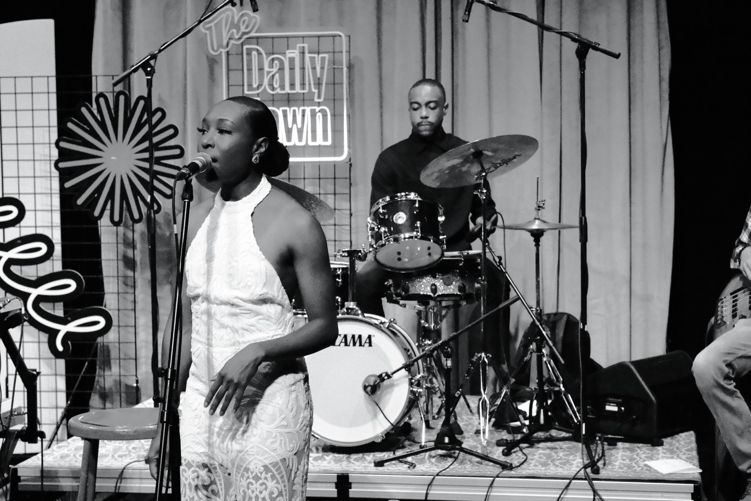 A black-and-white photo of Jasmine Cassell singing on the set of The Daily Jawn. The drummer is visible behind her.