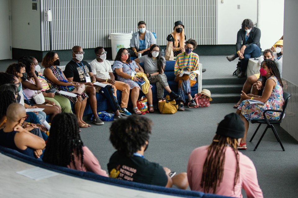 Workshop participants sit in a semi circle around a seated presenter. All are wearing masks inside.