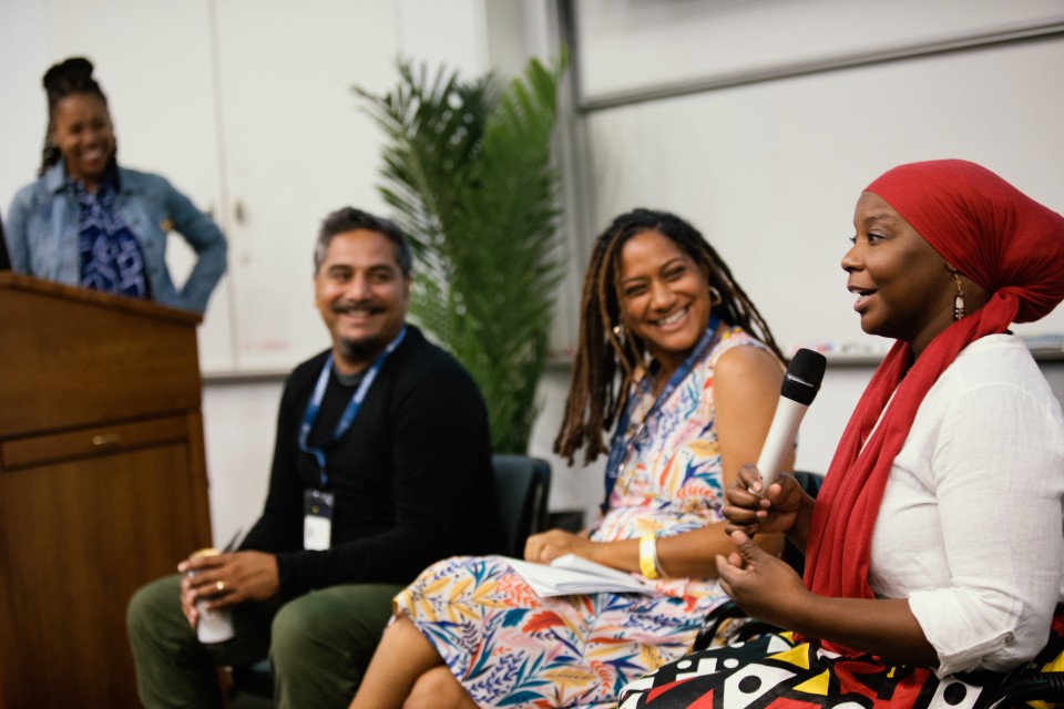 Sonya Childress sits in the middle of a row of three people. The person on the far right is holding a microphone. Sonya and the person to her left are looking at the person with the microphone and smiling.