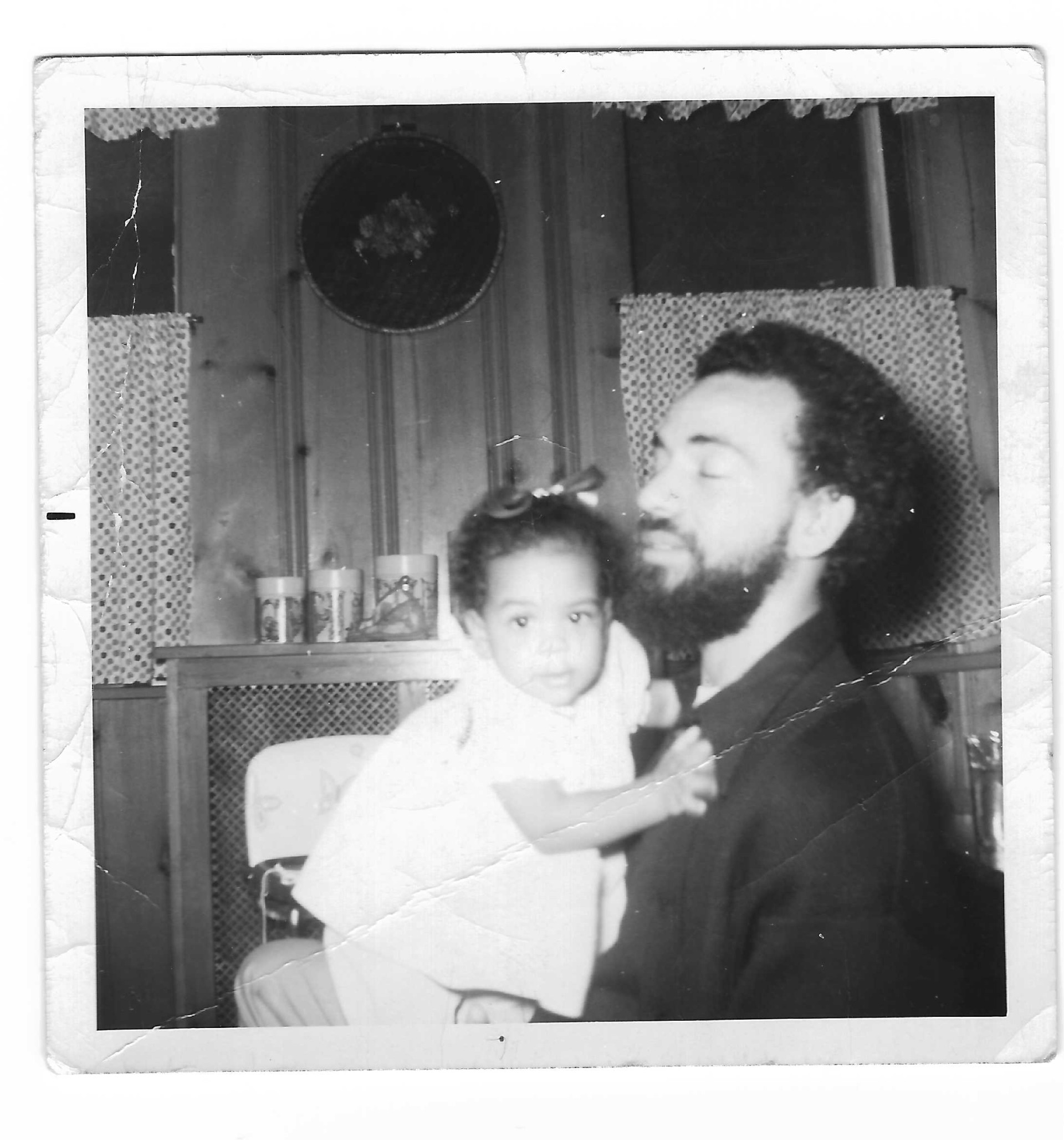 A black-and-white photo of a man sitting with a baby girl. The girl is wearing a white dress and has a bow in her hair. She is clinging to the man, who is presumably her father.