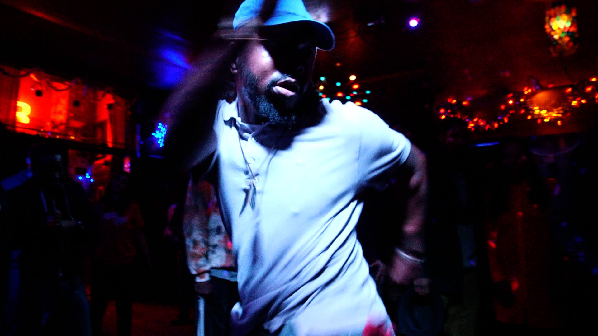 A photo of a Black person dancing at a club. They are wearing a white polo and a baseball cap. The photo is slightly blurry, there are colorful string lights glowing in the background.