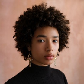 A headshot of Nile. She has an afro and gold earrings. Her expression is straight-faced and focused.