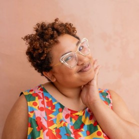 A headshot of Zoe Greggs, they have light brown skin, short curly hair, and large gold-rimmed glasses. They are smiling and tilting their head to the left.