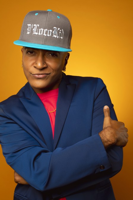 A headshot photo of D'Lo. He is a queer Tamil-Sri Lankian-American person. He is wearing a hat, a blue blazer, and a red shirt. He has a smirk on his face.