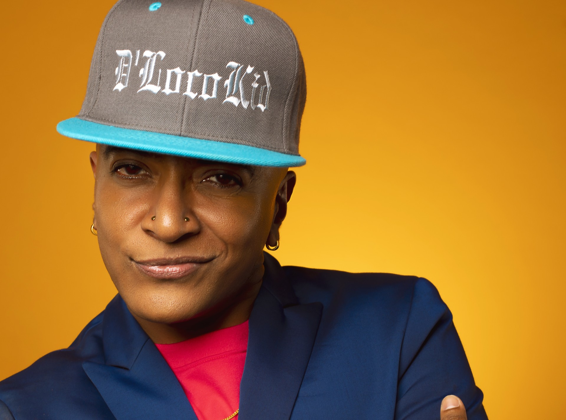 A headshot photo of D'Lo. He is a queer Tamil-Sri Lankian-American person. He is wearing a hat, a blue blazer, and a red shirt. He has a smirk on his face.