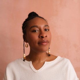 A headshot of Akua Maat. She is wearing a white shirt and in front of a salmon colored backdrop, looking directly at the camera.