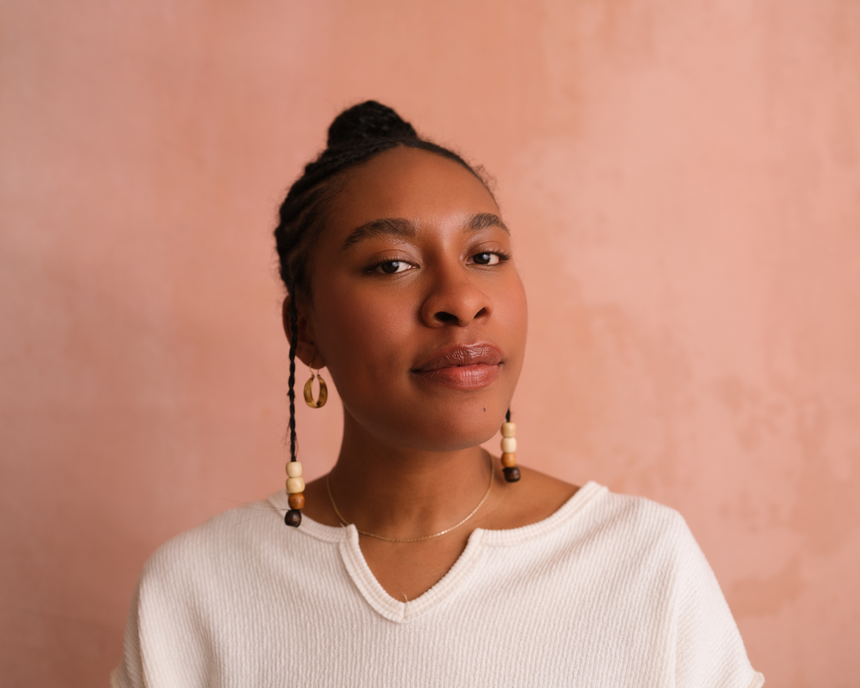 A headshot of Akua Maat. She is wearing a white shirt and in front of a salmon colored backdrop, looking directly at the camera.