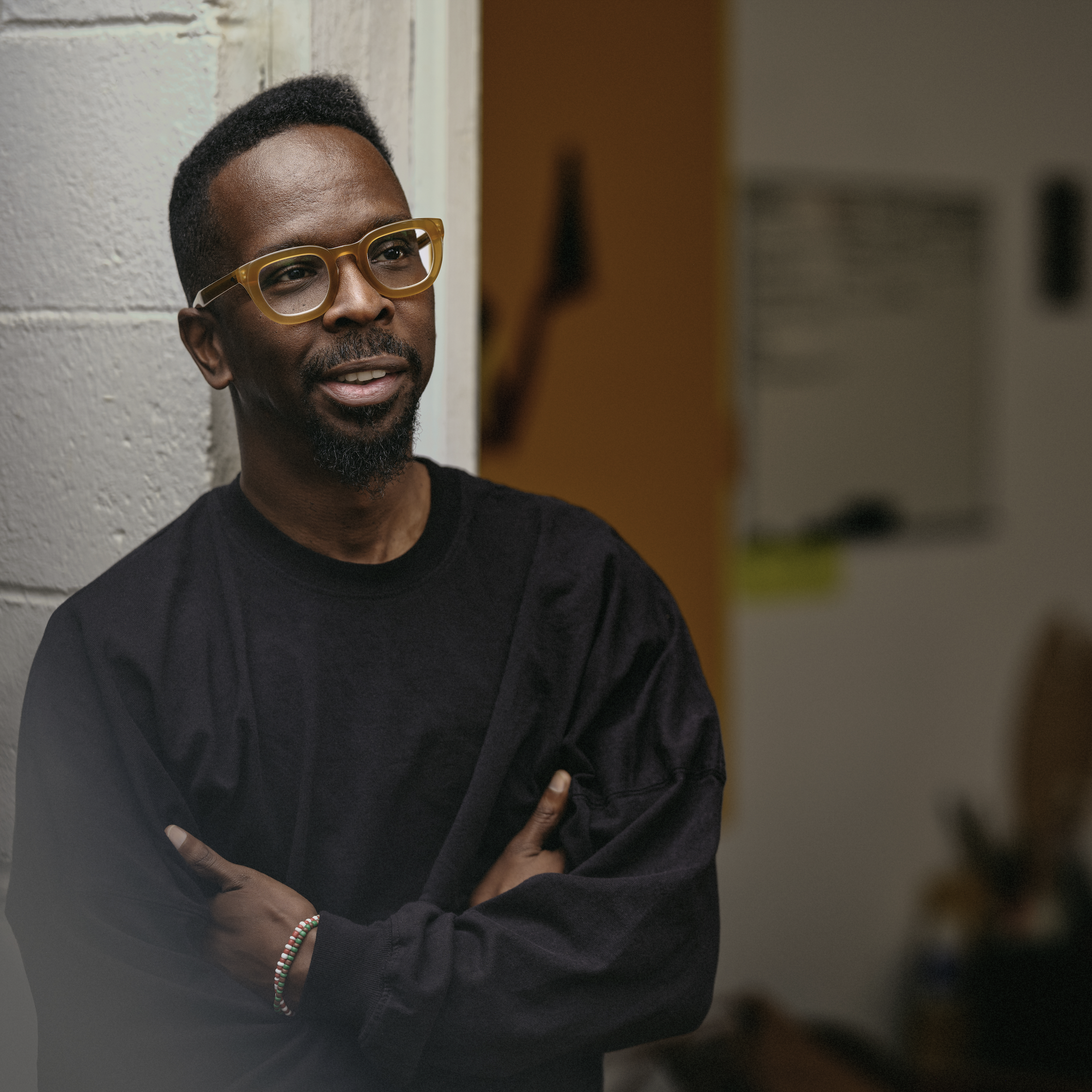 An image show a portrait of Dr. Fahamu Pecou leaning his back against one side of an entryway of what appears to be a classroom. His arms are crossed and his hands are tucked underneath his arms. He is looking at something outside of the frame.