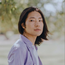 A picture of a non-binary person named Zhou Hao. He has long, straight black hair that frames his face and slightly falls towards his shoulders. He is wearing a light purple shirt over a white top. The background is softly blurred, comprising natural elements such as trees and greenery. His expression is neutral, yet slightly contemplative, with his gaze directed slightly off-camera.
