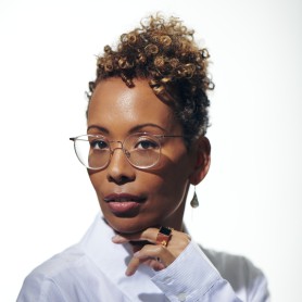 A close-up photo of J. Wortham, a Black person with short curly hair, wearing glasses and an earring in their right ear. Their head is slightly tilted to the left as they look at the camera.