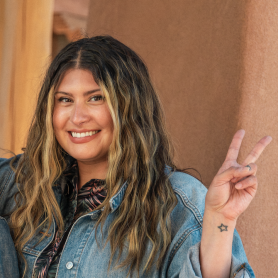 A picture of a woman named Alexandra lazarowich. She is posing next to an adobe wall with a wide smile on her face. She is making a peace sign with her right hand, which has a small star tattoo on the wrist. Alexandra's hair is long, wavy, and dark with streaks of light blonde. She is wearing a denim jacket over a patterned shirt.