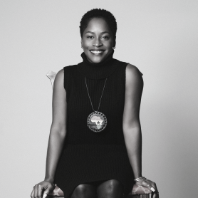 A picture of a woman named Yaba Blay. The portrait is in black-and-white and depicts her seated on a wooden chair against a plain, light-colored background. She has short, natural hair and is smiling warmly at the camera. She is wearing a sleeveless, high-neck, ribbed dress, and a large, circular necklace with a decorative design in the center and her hands gently resting on the sides of the chair.