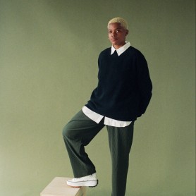 A picture of a non-binary person named Imani Dennison. They are standing against a soft green background. They have short, curly blond hair and are wearing a black sweater layered over a white collared shirt. Their outfit also includes dark green, loose-fitting trousers, and white sneakers with thick soles and tan accents. They have a calm and confident expression on their face with one of their feet casually propped up on a light wooden box with two handles on its sides, adding a sense of relaxed poise to their stance.