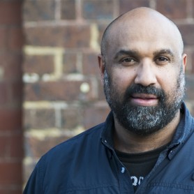 A picture of a man named John Harvey against a brick wall. He is bald with a short, graying beard and he is wearing a dark navy-blue jacket over a black shirt with white text, partially obscured. His expression is calm and slightly serious, with his eyes looking directly at the camera.