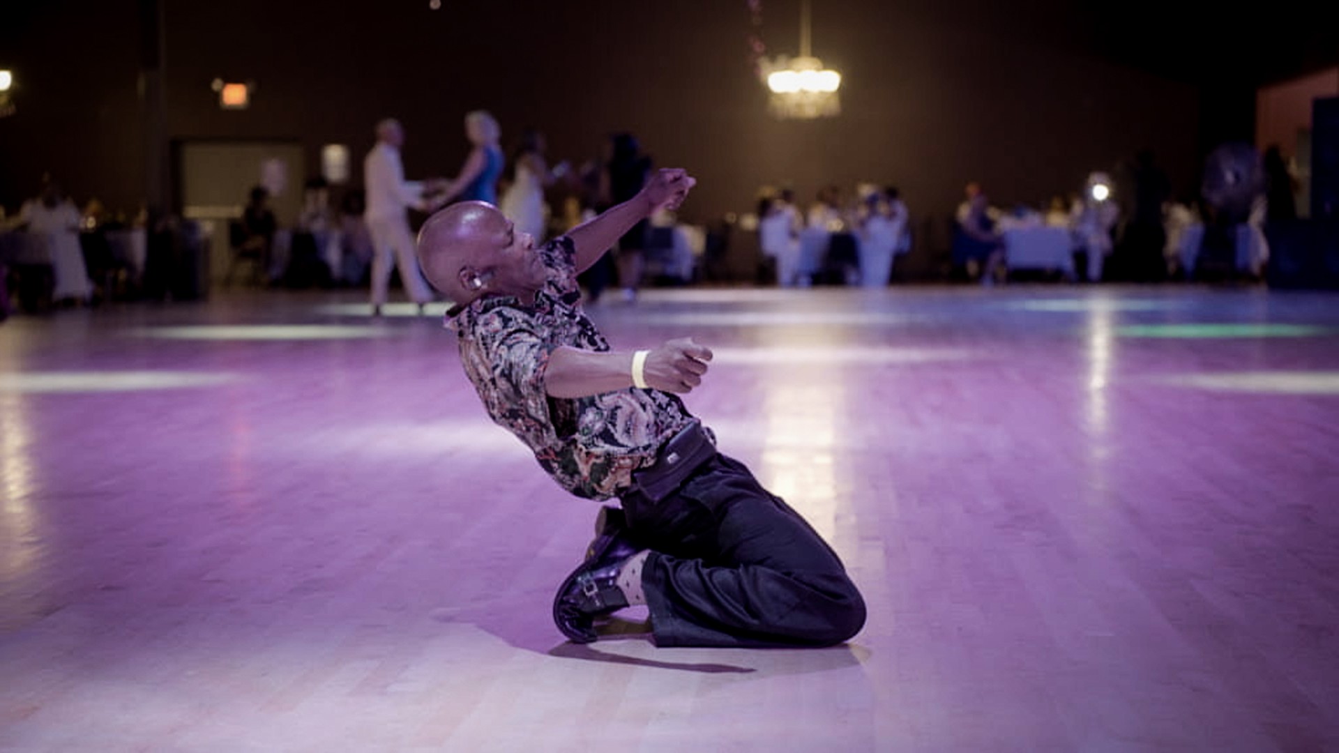 A photo of a dance hall. The edges of the room are lined with tables, there are a few couples dancing in the back. In the center there is a Black man, he is on his knees and appears to be mid-dance move. His arms are outstretched and his eyes are closed.