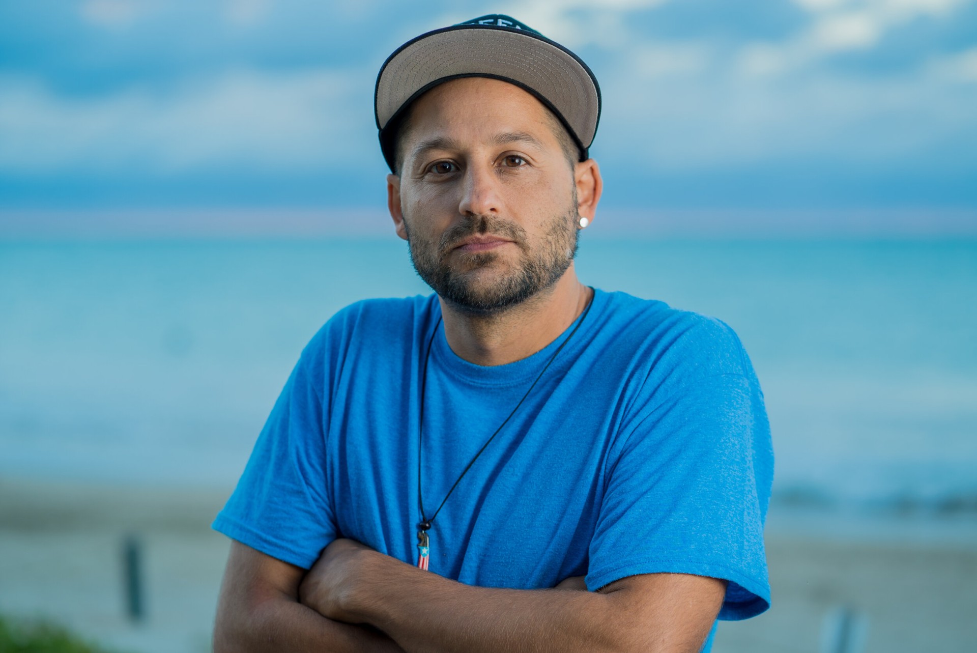 A headshot of Eli Jacobs-Fantauzzi, a Puerto Rican man. He is wearing a blue shirt and a baseball cap.
