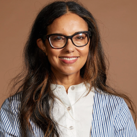 A headshot of Heidi Saman. She is brown woman. She is wearing a button-down shirt, a striped blazer, and glasses. She has long brown hair.