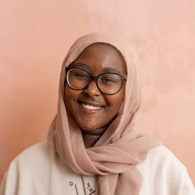 A headshot of Amina. She is a young Black woman. She is wearing a hijab and glasses. She is smiling at the camera.