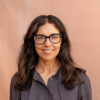 A headshot of Heidi Saman. She is brown woman. She is wearing a button-down shirt and glasses. She has long brown hair.