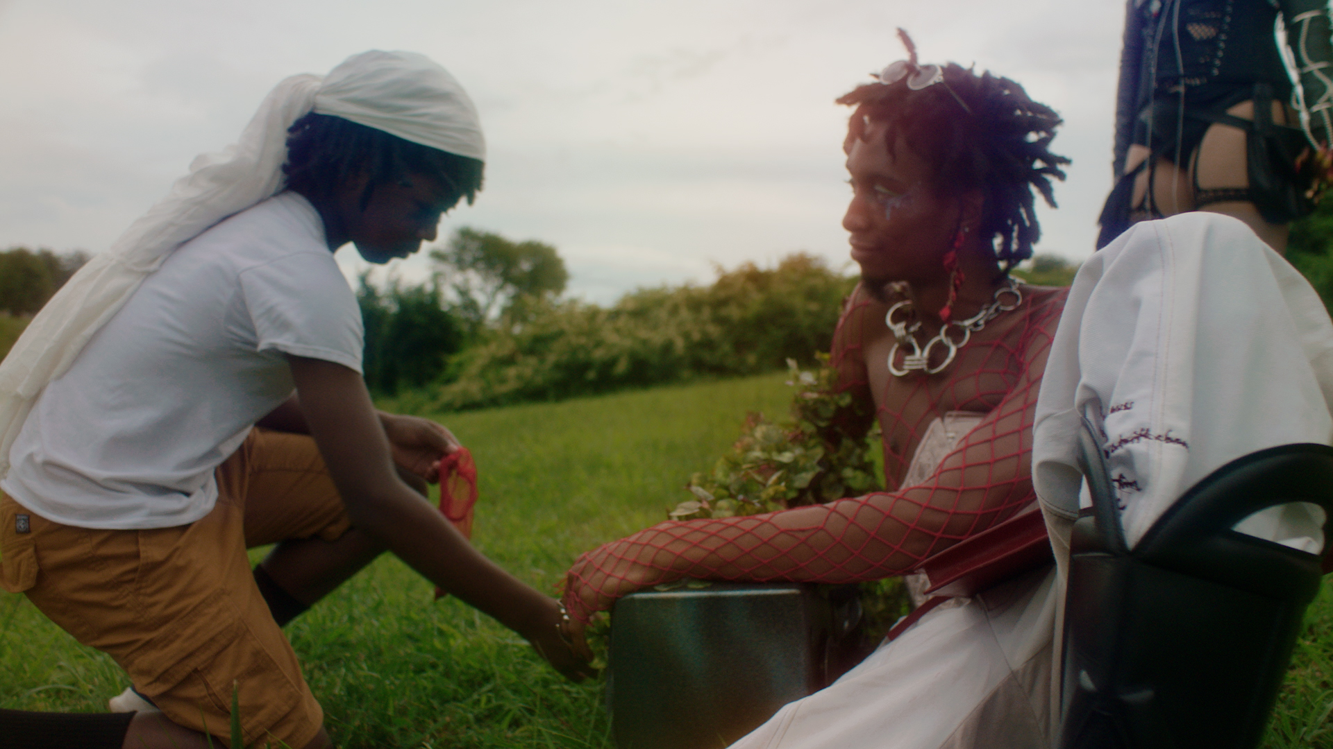 A still from One Magenta Afternoon. It shows a young Black boy sitting in a field with an older Black person. The older person has a soft expression on their face, the younger is looking at them with intrigue.
