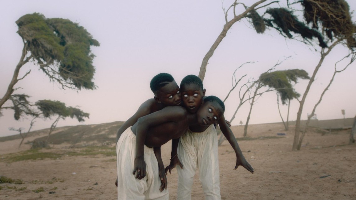 A film still shows three Black boys standing in a desert, their bodies intertwined as they stare at the camera with white eyes.