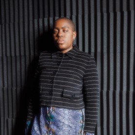 A picture of a woman named Onyeka Igwe leaned towards a black textured foam wall. Their hair is buzzed, and they are wearing a black and white horizontal striped collared long sleeve shirt. One of their eyes is faced forward and the other is almost closed, and they have a serious expression.