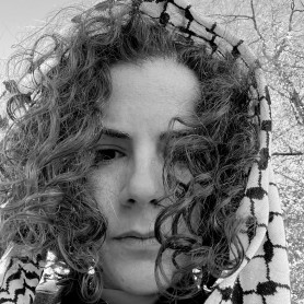 A black-and-white picture of a genderqueer person with curly hair named Razan AlSalah. They are looking down towards the camera with blank expressions, several strands of hair covering one of their eyes. Atop their head draped is a patterned scarf called a keffiyeh, with branches and a blank sky peeking out from the background.