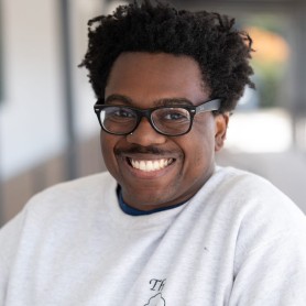 A picture of a man named Spencer Garland. He smiles broadly against a blurred outdoor background wearing a light gray crewneck and black glasses. He has short curly hair, a light mustache, and a stud nose ring.