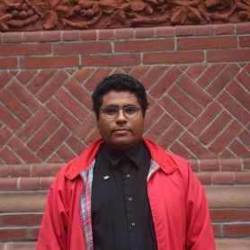 A picture of a man named Luis Arnias. He is wearing round army green glasses, and an open bright red coat lined with plaid inside. Underneath the coat is a black button down matching his jet-black short hair. He is against a geometrical brick background and is staring straight at the camera with a neutral expression.