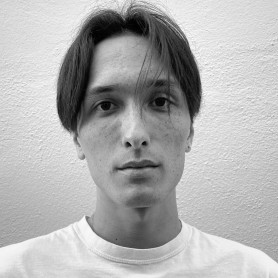 A black-and-white picture of a man named Devin Jie Allen against a light plain wall. He is wearing a plain white t-shirt and a resting facial expression. Several strands of his hair gently rest on his face, framing one of his eyes looking deeply into the camera.