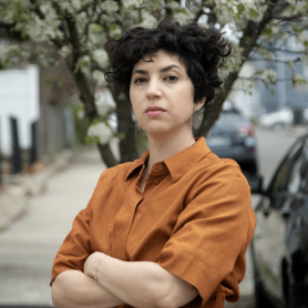A picture of a woman named Mary Jirmanus. She is wearing a burnt orange blouse with her arms crossed against an outdoor background of a blooming tree. She is turned slightly but staring at the camera with fierce eyes and short black curly hair.
